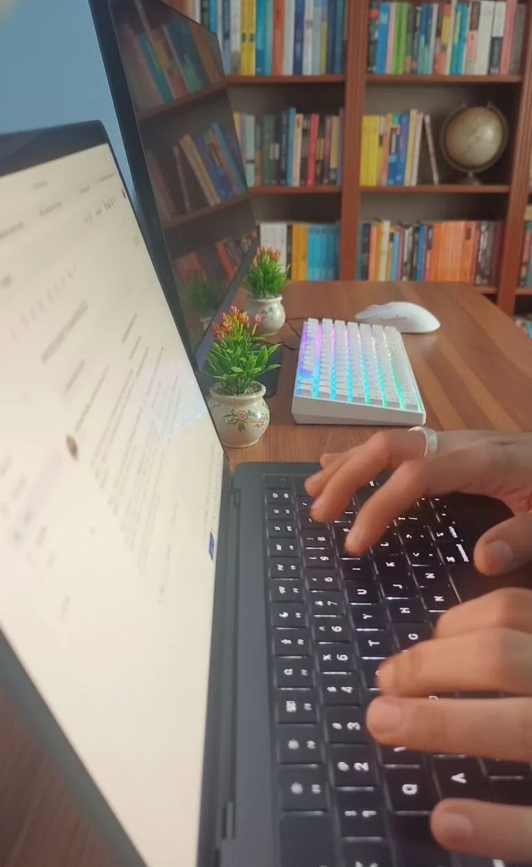 Hands typing on a laptop at a desk, a wall of bookshelves behind