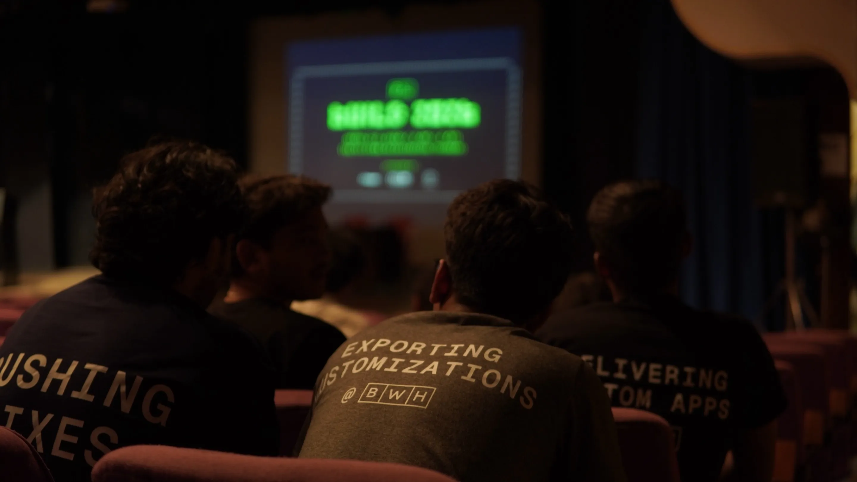 Four people in BWH tees watching a talk from the back rows of a dark auditorium